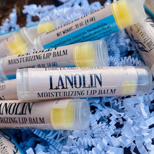 May include: A close-up of several tubes of lanolin moisturizing lip balm. The tubes are clear with a white label that says "Lanolin Moisturizing Lip Balm". The tubes are sitting on a bed of blue and white crinkled paper.