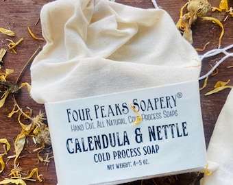 Könnte beinhalten: Ein Stück kaltverseifte Seife in einem cremefarbenen Stoffbeutel mit weißem Papieretikett. Das Etikett trägt die Aufschrift Four Peaks Soapery, Hand Cut, All Natural, Cold Process Soaps, Calendula & Nettle, Cold Process Soap, Nettogewicht: 113-142 g.