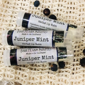 May include: Three tubes of lip balm with a white background. The tubes are labeled "Juniper Mint Herbal Lip Balm" and "Four Peaks Soapery Made in the Southwest".