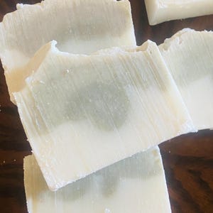 May include: Close-up of several handmade soap bars. The bars have a layered design with a white top and a light green base. The soap has a textured surface and a natural, rustic appearance.