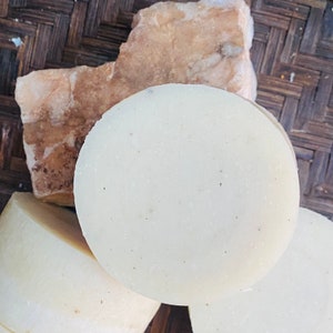 May include: Three round, white, handmade soap bars with a speckled texture. The soap bars are on a woven brown surface.