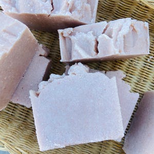 May include: Several rectangular bars of light pink soap with a textured, uneven surface. The soap bars have a matte finish and appear to be handmade, showing slight variations in shape and size across the collection of stacked pieces.
