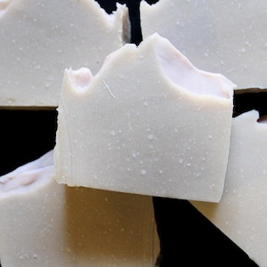 May include: Close-up of several bars of handmade soap. The soap is a pale, creamy color with a slightly textured surface and small white droplets. The edges of the soap have a rough, uneven appearance. The soap appears to be stacked.