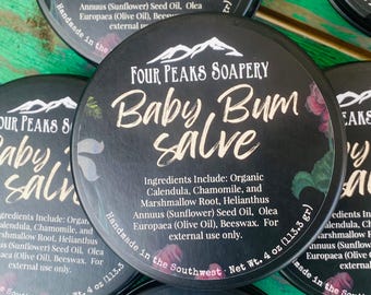 May include: Several black circular containers of baby bum salve are displayed. The containers feature the text "Four Peaks Soapery" and "Baby Bum Salve" in white script. Ingredients include organic calendula, chamomile, and sunflower seed oil. Each container is 4 oz.