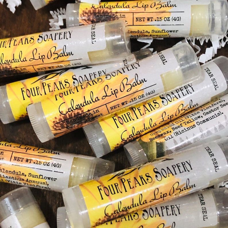 以下が含まれることがあります： Four Peaks Soaperyの「Calendula Lip Balm」のリップバームチューブのコレクション。各チューブはひまわりのグラフィックで「Calendula Lip Balm」と表示されています。チューブは透明で、中のバームが見え、内容量は4gです。