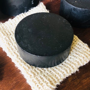 May include: A black circular bar of soap resting on a natural fiber bath sponge. The soap is smooth and has a matte finish.