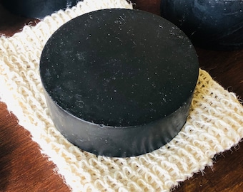 May include: A black circular bar of soap resting on a natural fiber bath sponge. The soap is smooth and has a matte finish.