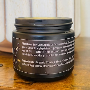 May include: A black jar with a screw-on lid. The label includes text about directions for use and ingredients, including organic rosehip and beeswax. The jar is on a wooden surface.