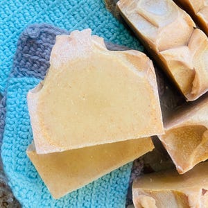 May include: Close-up of several bars of handmade soap with a light yellow hue and speckled texture. The soap is resting on a blue and gray crocheted washcloth. The soap has a rustic, natural appearance.