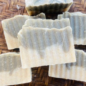 May include: Several handmade soap bars with a two-tone design. The top portion is a muted green, while the bottom is a creamy beige. The bars are rectangular with a slightly textured surface, suggesting a natural, artisanal quality.