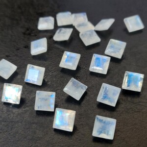 May include: A collection of 18 iridescent white moonstone square cabochons. The stones are approximately 10mm x 10mm in size and have a smooth, polished finish.