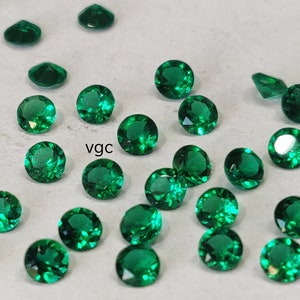 May include: A collection of small, round, faceted emerald green gemstones scattered across a surface. Each stone features a brilliant cut with multiple reflective surfaces. The letters vgc are printed in black in the center of the arrangement.