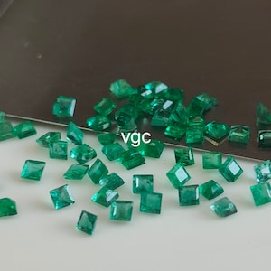 May include: A collection of emerald green square-shaped gemstones, scattered on a white surface. The gemstones are various sizes and have a polished, faceted finish.