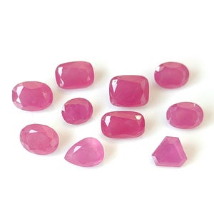 May include: Assortment of ten pink ruby gemstones in various shapes and sizes, including oval, pear, and rectangular cuts.