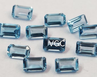 May include: A group of 11 light blue topaz gemstones, cut in an emerald shape. The gemstones are arranged in a random pattern on a white surface.