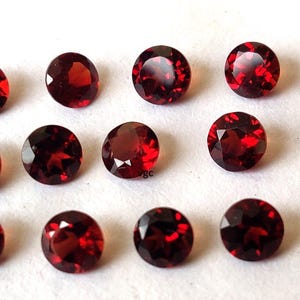 May include: A collection of fifteen round, faceted, deep red gemstones. The gems are arranged on a white surface, each displaying a brilliant, reflective quality. The stones are of similar size and shape, showcasing a consistent, polished appearance.