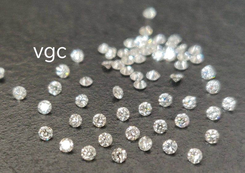 May include: A close-up image of a group of loose diamonds on a dark gray surface. The diamonds are round and brilliant, with a variety of sizes. The text "vgc" is visible in the upper left corner of the image.