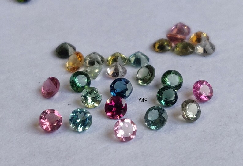 Puede incluir: Surtido de piedras preciosas redondas y facetadas en varios colores, incluyendo rosa, verde, azul y amarillo. Las piedras preciosas est&aacute;n dispuestas sobre una superficie blanca.