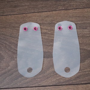May include: Two white plastic pieces with pink eyelets. Each piece has a rounded top and a hole at the bottom.