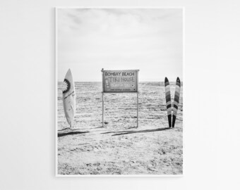 May include: Black and white photo of a beach with two surfboards and a sign that reads "Bombay Beach Tiki House Surf Ski".