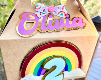 Könnte beinhalten: Eine braune Papp-Geschenkbox mit einem Regenbogen, Wolken und der Zahl 2. Die Box hat auch ein lila und goldenes Namensschild mit der Aufschrift "Olivia".