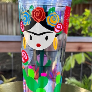 May include: A clear plastic tumbler with a green straw and a colorful design featuring a woman with flowers in her hair and cacti. The design is reminiscent of Frida Kahlo's artwork. The tumbler has the word "STARBUCKS" printed on the side.