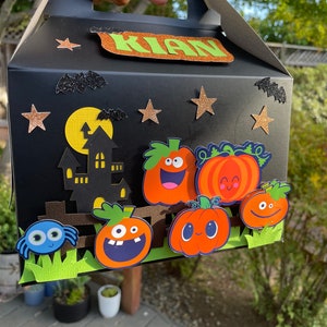 Può includere: Una scatola di dolcetti di Halloween nera con un'etichetta con il nome "Kian". La scatola è decorata con pipistrelli, stelle, una luna, una casa stregata e una recinzione con zucche e un ragno.