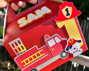 Peut inclure: Boîte à dragées de camion de pompiers rouge et jaune avec un chapeau de pompier et un chien portant un chapeau de pompier. La boîte porte l'inscription "SAM" et "1".
