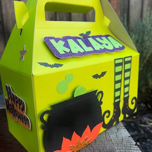 May include: A green and purple Halloween treat box with a cauldron, witch's legs, and bats. The box has a handle and says "Happy Halloween" and "KALAYA" on it.