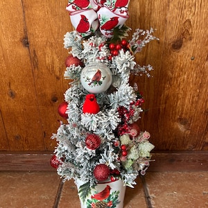 May include: A tabletop artificial Christmas tree with flocked branches in a white metal bucket. The tree is decorated with red ornaments, red berries, a small red bird figurine, and a white ornament with a cardinal and the text "When cardinals appear, angels are near". A large bow with a cardinal print sits at the top.