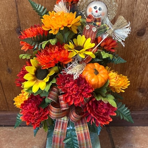 May include: A decorative autumn arrangement featuring a scarecrow with a straw hat, orange and red chrysanthemums, yellow sunflowers, a small orange pumpkin, and a plaid ribbon, all set into a green foam base with green foliage accents.