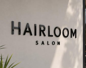 May include: A wall-mounted sign featuring the word HAIRLOOM in large, bold, black capital letters, with the word SALON positioned directly underneath in a smaller, matching black font, set against a smooth, light-colored, off-white wall surface.