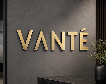 May include: A three-dimensional wall sign featuring the word VANTÉ in brushed gold metallic lettering. The letters are mounted on a dark gray textured wall surface, with a small green plant in a black vase visible in the lower right corner.