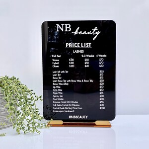 May include: Black acrylic sign with white text listing prices for beauty services, including lashes, waxing, and facials. The sign reads "NB beauty" and "#NBBEAUTY".