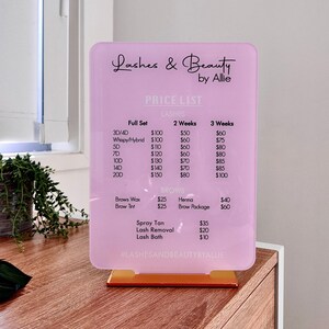 May include: Pink price list sign with a white border and gold stand. The sign lists prices for lashes and brows services, including full sets, 3D/4D, whispy/hybrid, 5D, 7D, 10D, 14D, 200, brows wax, brow tint, henna, brow package, spray tan, lash removal, and lash bath. The sign also includes the text "Lashes & Beauty by Allie" and "#LASHESANDBEAUTYBYALLIE".