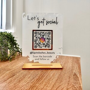 Lets get social sign with white splash background and gold mirror combination, QR code in the middle with Instagram logo, and instagram username under the QR code, and instruction to scan the Qr code to follow under the handle