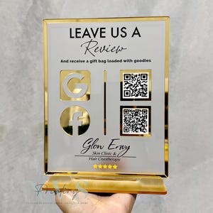 May include: A gold-framed acrylic sign with the text "LEAVE US A Review" and "And receive a gift bag loaded with goodies." Includes Google and Facebook logos, QR codes, and the business name "Glow Envy Skin Clinic & Hair Cryotherapy."