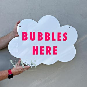 以下が含まれることがあります： 白い雲の形をしたサインで、明るいピンク色で「BUBBLES HERE」と書かれています。サインには吊り下げ用の穴が2つあります。楽しくて遊び心のある装飾品です。