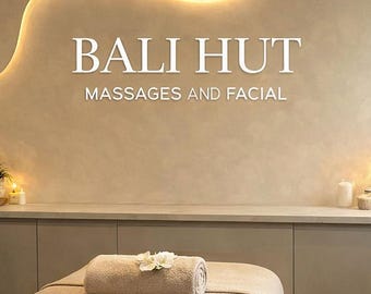 May include: Interior of a spa with the text "BALI HUT MASSAGES AND FACIAL" on the wall. A massage table is covered with a beige sheet and a rolled towel with a flower. Two wicker lamps and candles provide soft lighting.