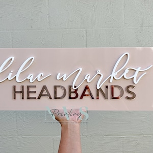 Rectangle acrylic business sign in blush pink acrylic backing installed on the wall, LILAC MARKET in 3D gloss white acrylic on top in cursive font, HEADBANDS under it in 3D rose gold mirror in SANS font