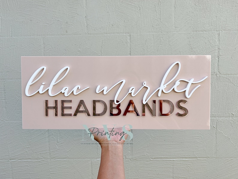 Rectangle acrylic business sign in blush pink acrylic backing installed on the wall, LILAC MARKET in 3D gloss white acrylic on top in cursive font, HEADBANDS under it in 3D rose gold mirror in SANS font
