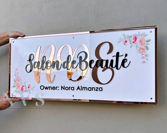 May include: A white sign with a rose gold frame and black lettering that reads "Salon de Beauté" with the owner's name "Nora Almanza" below. The sign has a floral design in the top right corner.