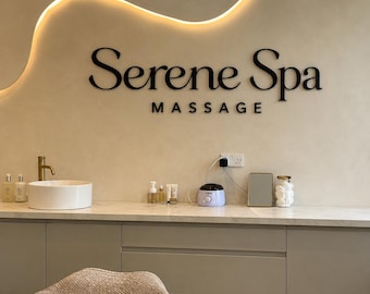May include: Interior shot of a spa with the words "Serene Spa MASSAGE" in black lettering on a beige wall. A white sink with a gold faucet is visible, along with various spa products and a wax warmer.