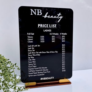 May include: A black acrylic sign with white text listing prices for beauty services, including lashes, lash lifts, tinting, waxing, facials, and spray tans. The sign is on a gold stand and reads "NB beauty PRICE LIST" at the top.