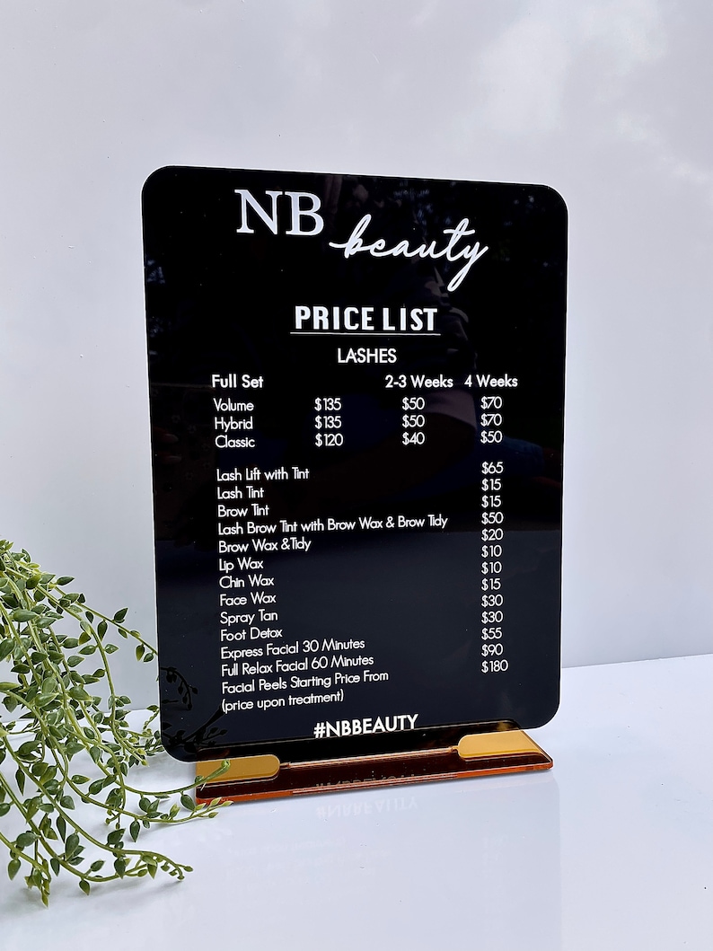 May include: A black acrylic sign with white text listing prices for beauty services, including lashes, lash lifts, tinting, waxing, facials, and spray tans. The sign is on a gold stand and reads "NB beauty PRICE LIST" at the top.