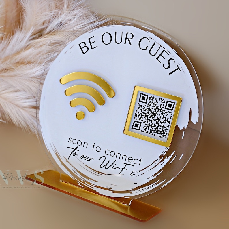 Op de afbeelding: Rond wit acryl bord met gouden accenten. Het bord zegt "Be Our Guest" en heeft een wifi-symbool en een QR-code. De tekst onder de symbolen zegt "Scan to connect to our Wi-Fi".