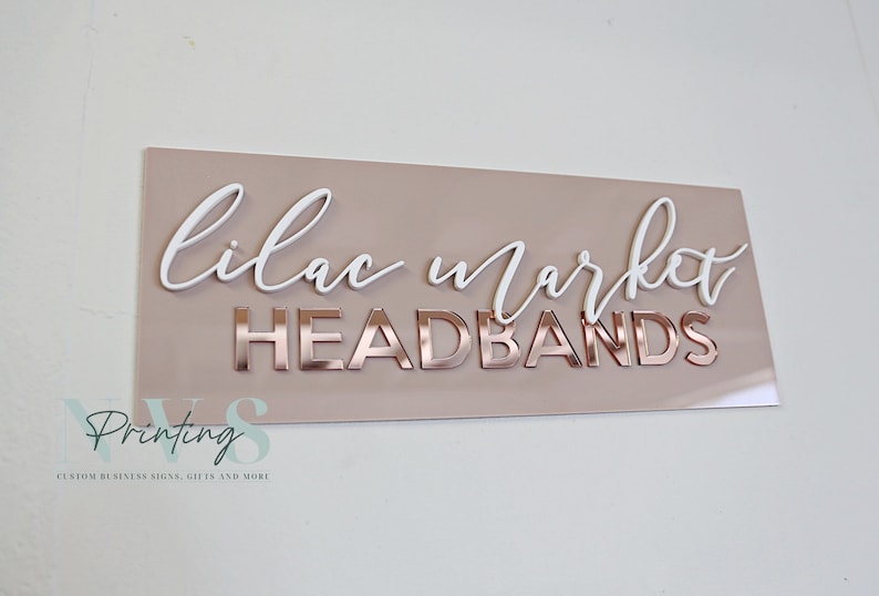 Rectangle acrylic business sign in blush pink acrylic backing installed on the wall, LILAC MARKET in 3D gloss white acrylic on top in cursive font, HEADBANDS under it in 3D rose gold mirror in SANS font
