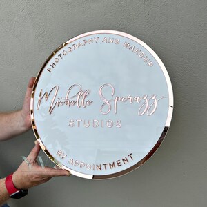 Acrylic Round Business Sign against the wall in white acrylic backing with rose gold mirror round border frame and 3D letters in rose gold mirror acrylic, photography and make up along the top and business studio name along the bottom