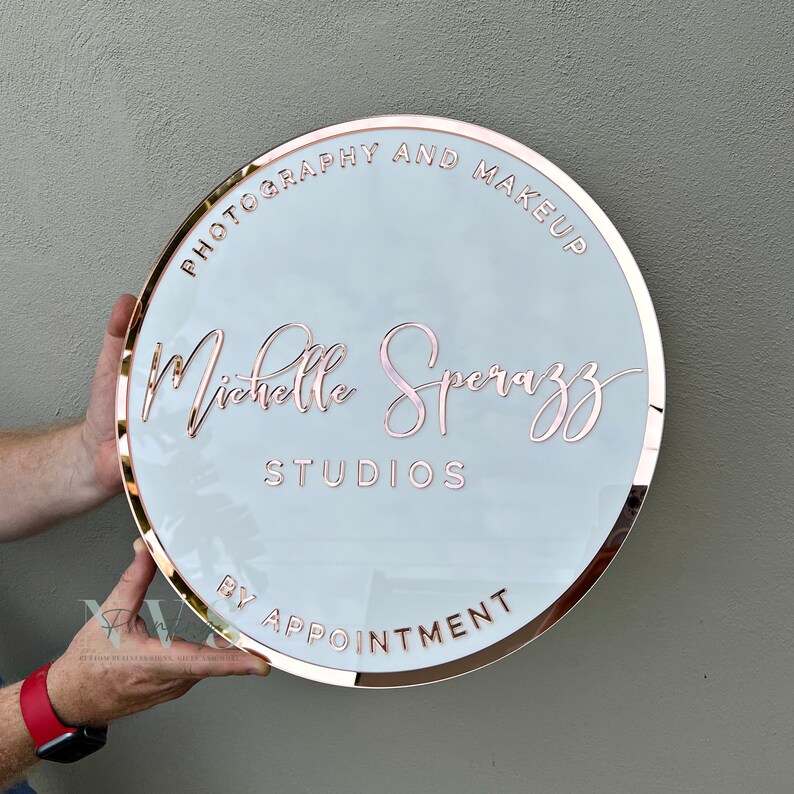 Acrylic Round Business Sign against the wall in white acrylic backing with rose gold mirror round border frame and 3D letters in rose gold mirror acrylic, photography and make up along the top and business studio name along the bottom