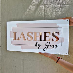 May include: A white sign with the words "LASHES by Jess" in rose gold and black lettering. The sign has a pink watercolor-style background and a black rectangular border. The sign is being held up by two hands.